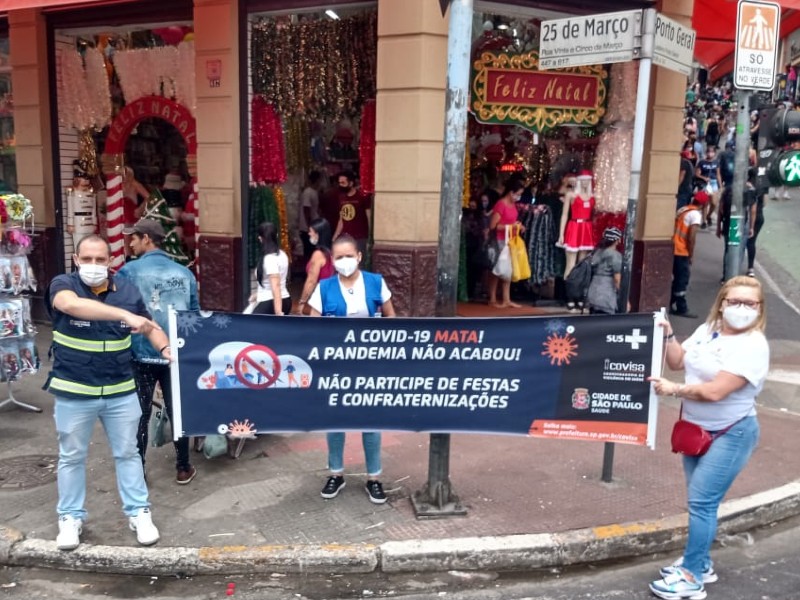 Um homem e duas mulheres seguram uma faixa com fundo azul em que está escrito "A Covid-19 mata! A pandemia não acabou. Não participe de festas e confraternizações". Eles estão em frente a uma loja de artigos natalinos na Rua 25 de Março